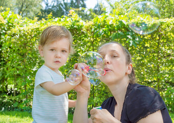 Obraz premium Cute little boy and his mother having fun blowing soap bubbles in the park. Funny outdoor activity for children in a beautiful summer sunny day