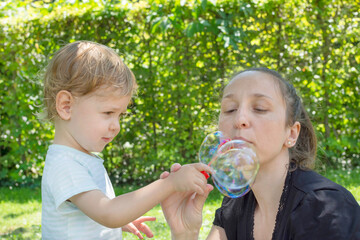 Cute little boy and his mother having fun blowing soap bubbles in the park. Funny outdoor activity for children in a beautiful summer sunny day