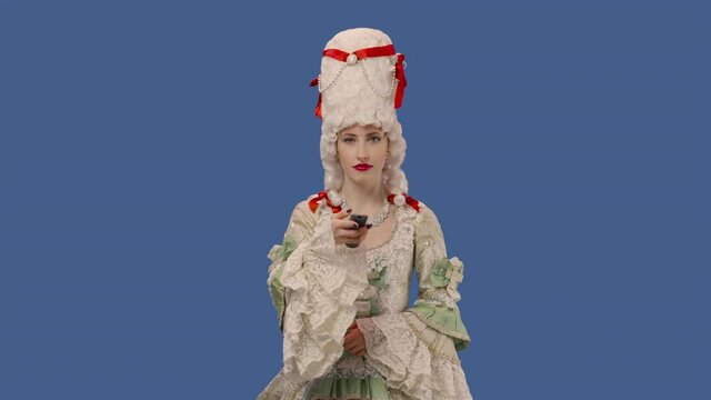 Portrait of courtier lady in white vintage dress and wig watching TV and changing channels using remote. Young woman posing in studio with blue screen background. Close up. Slow motion ready 59.94fps.