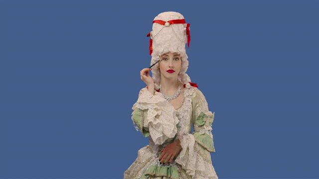 Portrait of courtier lady in white vintage lace dress and wig is doing makeup with a brush. Young woman posing in studio with blue screen background. Close up. Slow motion ready 59.94fps.