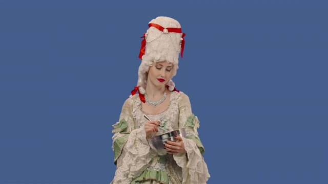 Portrait of courtier lady in vintage lace dress and wig is preparing food, whisking something in a bowl. Young woman posing in studio with blue screen background. Close up. Slow motion ready 59.94fps.
