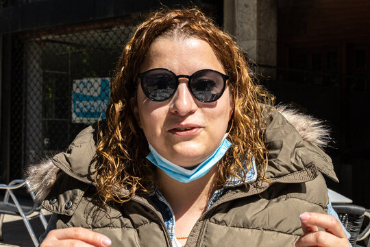 Curly-haired Woman Sitting On The Terrace Of A Restaurant Bar With Sunglasses And A Face Mask Pulled Down While Having Breakfast