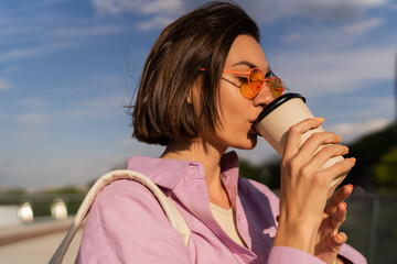 Summer portrait of pretty short haired woman in stylish sunglasses  enjoying coffee walking outdoor in sunny day. Wearing purple oversize shirt.