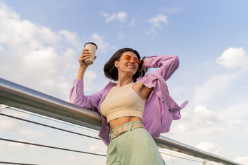 Happy woman with perfect figure  in stylish purple oversize shirt enjoying  cup of coffee  while walking on modern bridge.