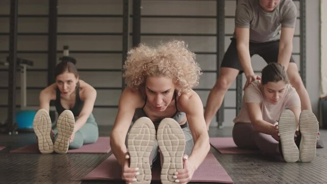 Full shot of three slim Caucasian women sitting on yoga mats in gym, doing seated forward bend, male fitness coach helping them