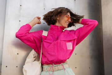 Fototapeta na wymiar Playful woman having fun in neon pink jacket over grey metallic urban wall. Fashionable sportive outfit.