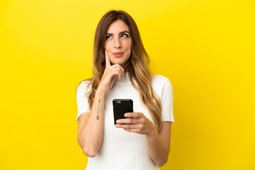 Caucasian woman isolated on yellow background using mobile phone and thinking