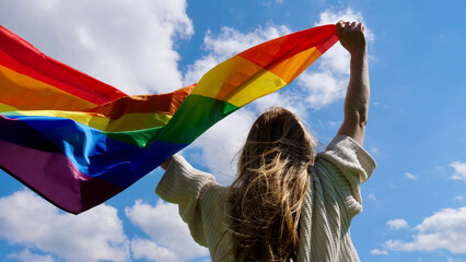 Blonde, lesbian, woman holding a rainbow LGBT gender identity flag on sky background with clouds on...