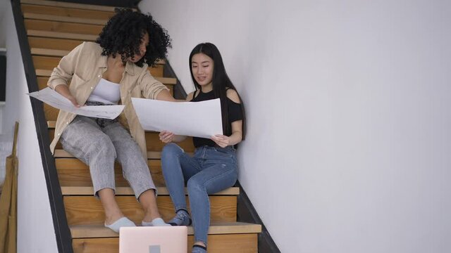 Two nervous young women arguing discussing project in home office. Portrait of confident worried Asian and African American startupers planning business idea sitting on stairs talking