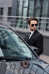 Fototapeta na wymiar bearded bodyguard in suit and sunglasses with security earpiece near modern auto