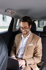 Fototapeta na wymiar cheerful businessman in glasses typing on laptop in car