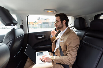 businessman in glasses working remotely with laptop while talking on cellphone in car