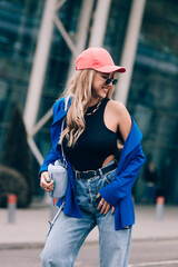 Young sexy blonde hipster woman posing on the street. Wearing blue stylish jacket, jeans and baseball hat and sunglasses.