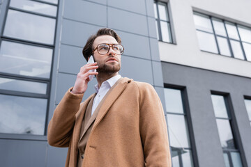 low angle view of man in glasses and beige coat talking on cellphone near building outside