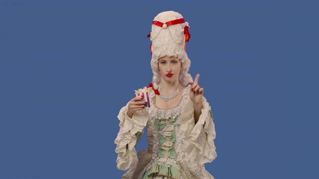 Portrait of courtier lady in vintage lace dress and wig, holding fries and showing thumbs down dislike. Young woman posing in studio with blue screen background. Close up. Slow motion ready 59.94fps.