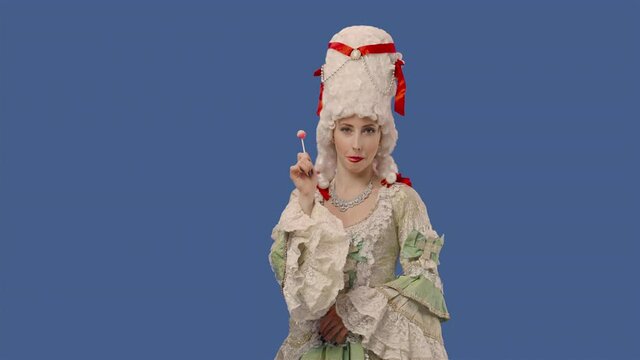 Portrait of courtier lady in white vintage lace dress and wig, enjoying sweet candy lollipop. Young woman posing in studio with blue screen background. Close up. Slow motion ready 59.94fps.
