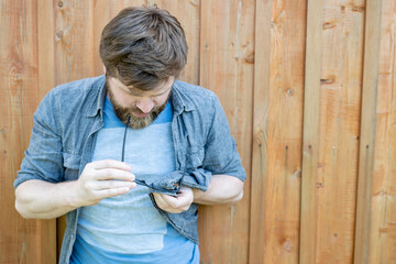 Concentrated man carefully cleans glasses from dust, wiping them with a shirt, against a background of a wooden wall. Copy space.