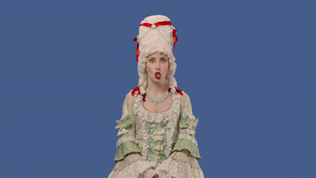 Portrait of courtier lady in white vintage lace dress and wig is chewing gum and blowing bubbles. Young woman posing in studio with blue screen background. Close up. Slow motion ready 59.94fps.