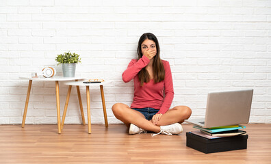 Young woman with a laptop sitting on the floor at indoors covering mouth with hand