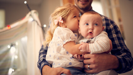 Close Up Of Young Girl Cuddling Baby Brother Whilst Sitting On Father's Knee At Home