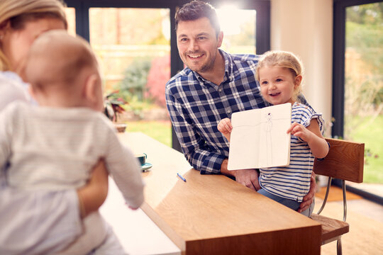 Family Sitting Around Kitchen Counter With Daughter Showing Parent Picture She Has Drawn