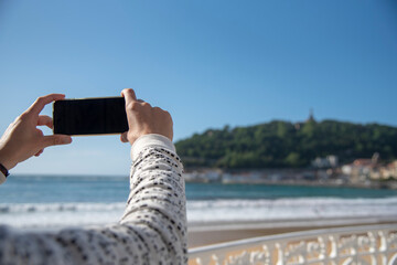 vista de las manos de hombre sacando una foto con un móvil a la playa de la concha de san...