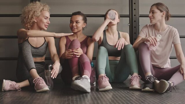 Full shot of four lean Mixed-Race and Caucasian sportswomen leaning on wall sitting on floor in gym, drinking water from bottles, talking and smiling after hard training