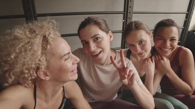 Handheld POV of four young Caucasian and Mixed-Race sports women sitting on floor in gym, smiling, talking, filming them on camera after training