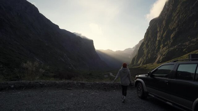 Girl getting out of her four wheel drive to take a picture of a deep scenic valley with the sun beaming through the mountains during sunset on to a cliff face in New Zealand.