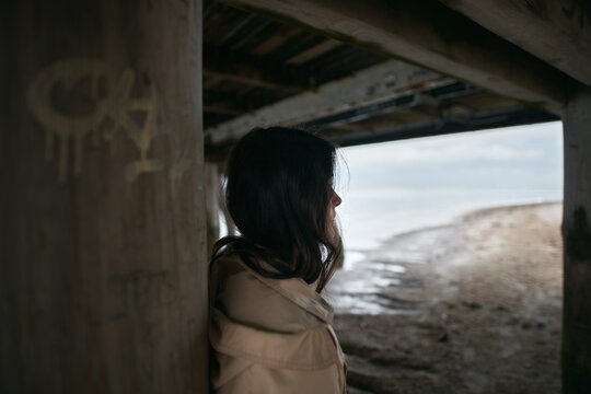 Portrait Of Stylish Brunette Under The Wooden Pier. Attractive And Trendy Girl With Long Brown Hair By The Seaside Stand In Tan Color Trench Coat.