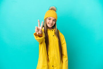Little girl wearing a rainproof coat over isolated blue background smiling and showing victory sign