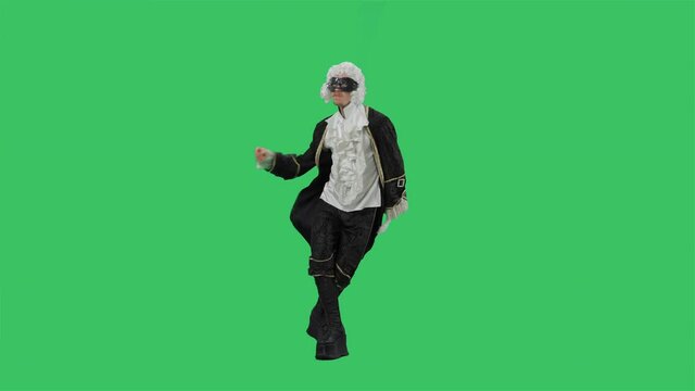 Portrait of courtier gentleman in black vintage suit, white wig and the mask, dancing merrily with fan in his hands. Young man posing in studio with green screen background. Full lenght. Slow motion.