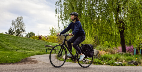 Fototapeta premium Adult Caucasian Woman Riding a Bicycle on a path in a modern city park. Spring Evening. Taken in Hawthorne Rotary Park, Surrey, Vancouver, British Columbia, Canada.