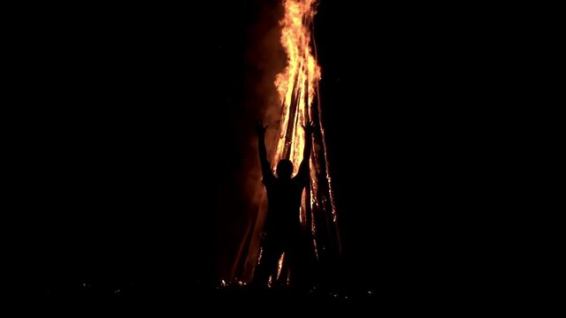 A man standing in the darkness in front of a big bonfire is holding his hands up and praying on it in slow motion. Wizard is making ritual with huge campfire at night. Concept of witchcraft and voodoo