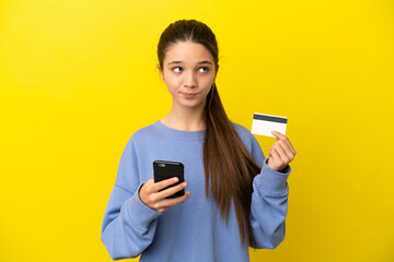 Little girl over isolated yellow background buying with the mobile with a credit card while thinking