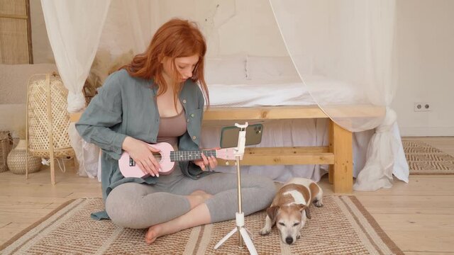 Girl playing ukulele at home in bedroom sitting floor with small cute dog Jack Russell terrier. Beautiful young long red haired girl in blue shirt chilling weekend mood. Video footage online tutorials