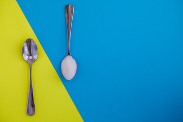 Sugar-replacement tablet of stevia and sugar in tea spoons lying in opposite directions on bright bicolor diagonal blue and yellow paper background. Horizontal, copy space, flat lay top view