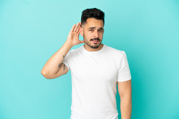 Young caucasian man isolated on blue background listening to something by putting hand on the ear