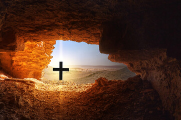 Black cross with bright light in front of dark cave in the dry hostile desert with boulders