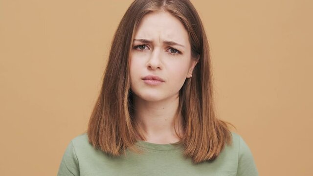 A serious young woman is shaking her head negatively while listening the viewer standing isolated over a beige wall in the studio