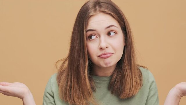 A confused young woman is shrugging shoulders standing isolated over a beige wall in the studio
