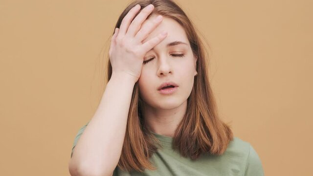 A displeased young woman is doing face palm gesture standing isolated over a beige wall in the studio