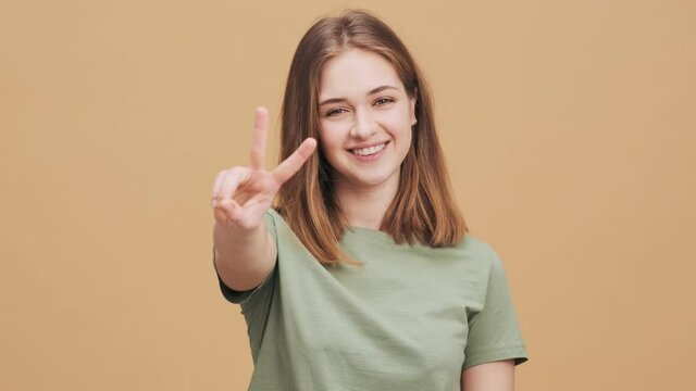A smiling young woman is doing peace gesture standing isolated over a beige wall in the studio