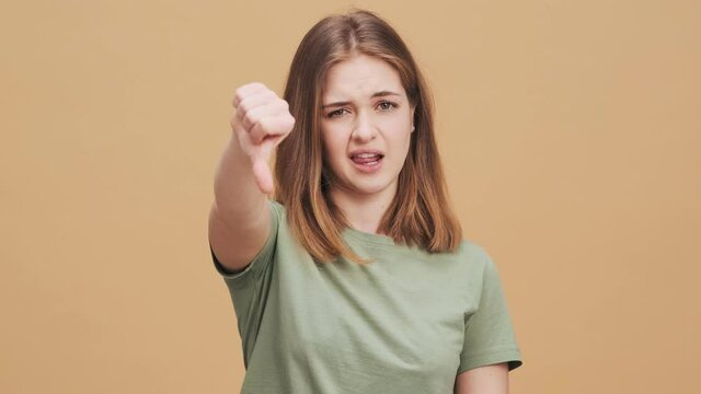 A displeased young woman is doing thumb-down gesture standing isolated over a beige wall in the studio