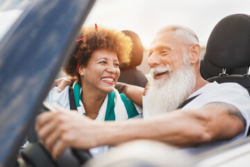 Senior couple having fun inside convertible car - Multiracial mature people on road trip in summer vacation with cabriolet car - obrazy, fototapety, plakaty