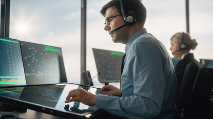Male Air Traffic Controller with Headset Talk on a Call in Airport Tower. Office Room is Full of Desktop Computer Displays with Navigation Screens, Airplane Departure and Arrival Data for the Team.