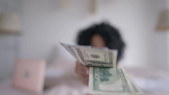 Rack focus from face of confident African American woman lying on bed to female hand holding dollars stretching to camera. Successful rich manager posing with cash income in home office indoors