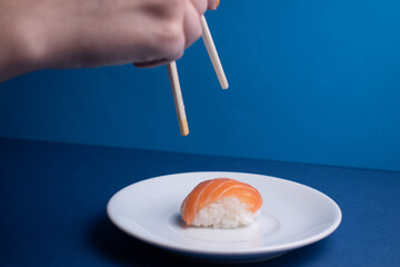 Hand of an unrecognizable person picking up sushi with chopsticks from a white plate on a blue background.