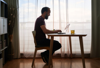 backlit portrait of middle-aged bearded man working with his laptop by the window of his home