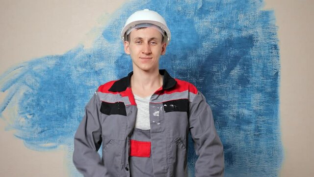 Jolly repairman in jumpsuit and protective construction helmet crosses hands in spacious apartment room against wall with blue paint spot closeup